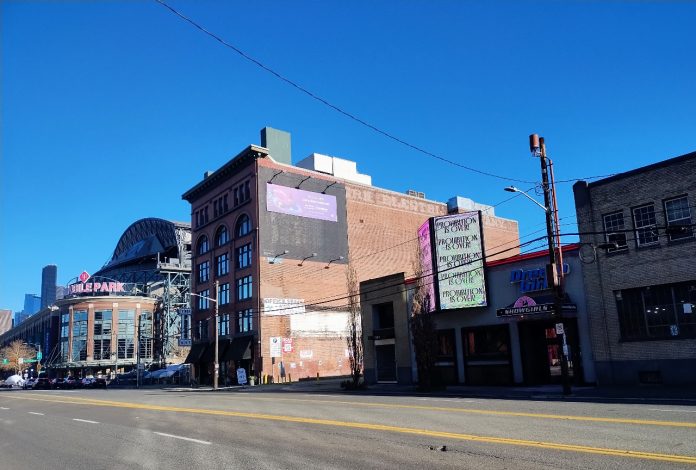 A photo from SoDo, with T-Mobile Park in the background. An older brick building stands next to smaller, more run down storefronts with a large wide road in the foreground