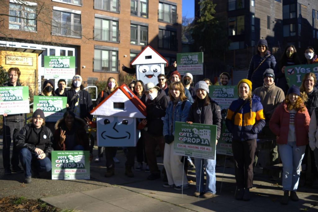 A group of about 20 poses in front of a brick apartment building, including folks in House-y mascot costumes.