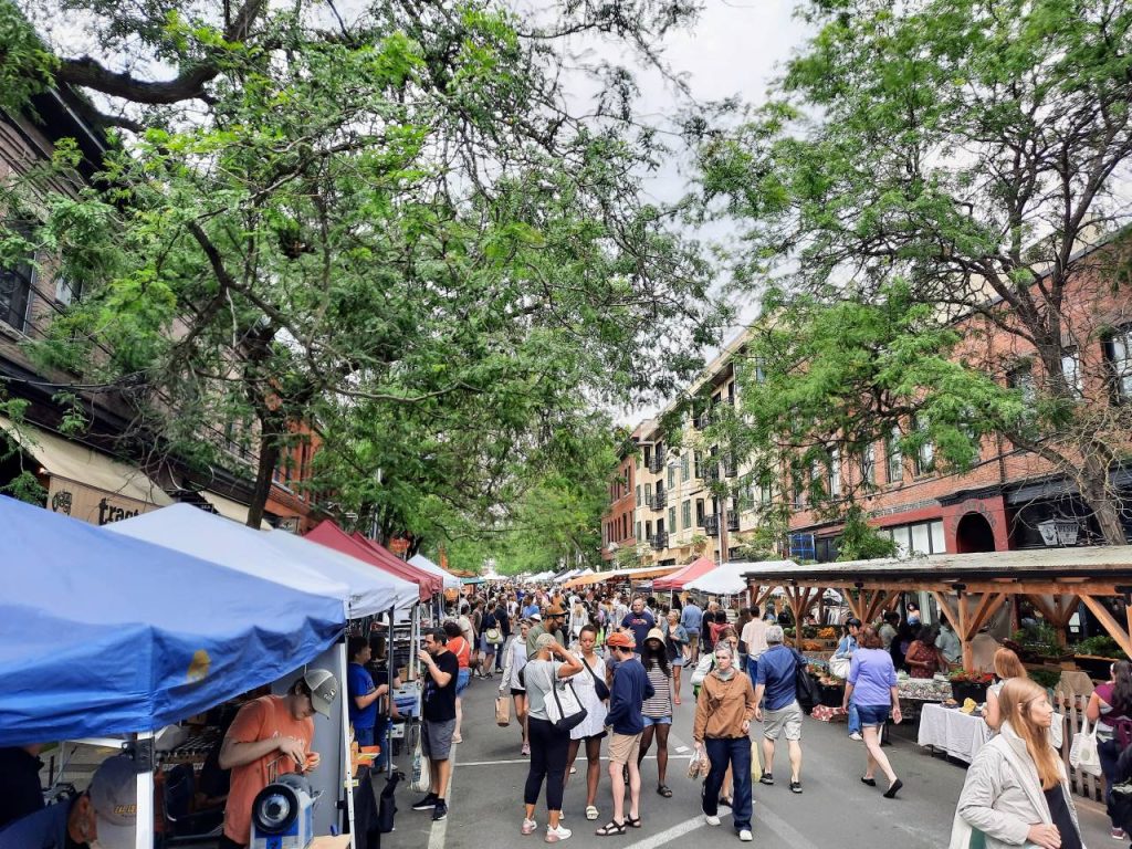 Farmers market stalls line the street and people fill the street, which is lined by trees. 