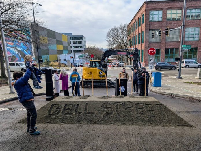 Bell Street is written into the pile of sand that was used in the ceremonial groundbreaking shoveling. A man hoists his child and officials mill about after the ceremony.