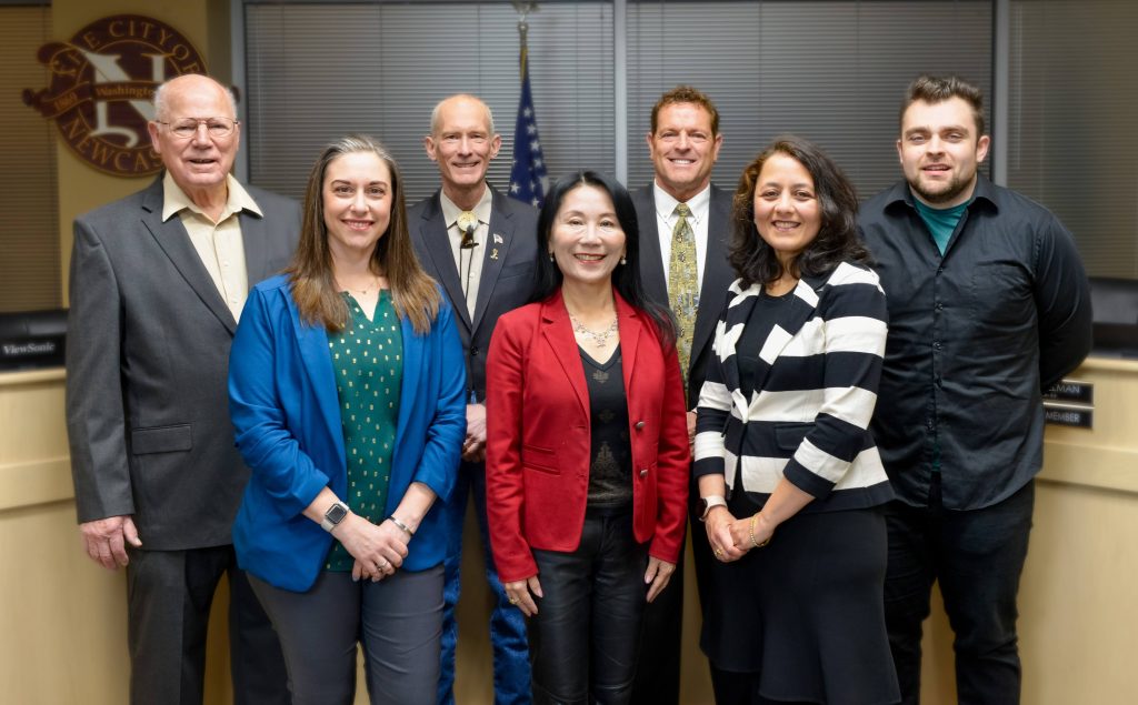 Clark is a bald white man wearing a bolo tie with his suit. He stands next to six city councilmembers in a group shot.