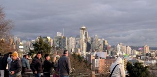 A handful of people stand at the railing at Kerry Park overlooking the Seattle skyline with the space needle prominently in the needle and they snap photos.