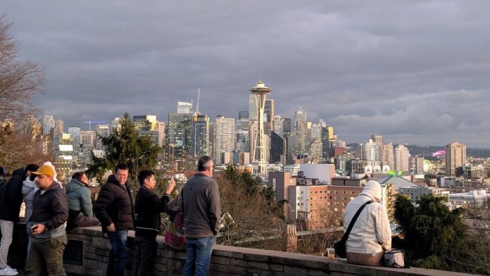 A handful of people stand at the railing at Kerry Park overlooking the Seattle skyline with the space needle prominently in the needle and they snap photos.