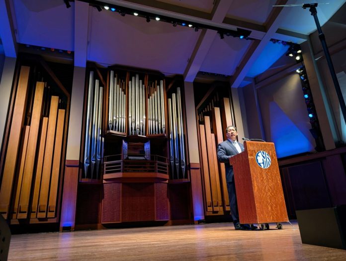 Harrell stands at a lectern with a big pipe organ behind him on the Benaroya auditorium stage.