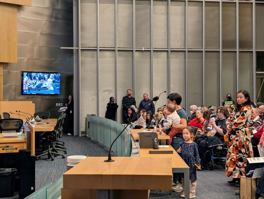 A women holds a baby with a toddler at her feet and speaks into the micro in Seatlte council chambers.