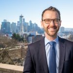 A white man in glasses and suit poses in from of the Seattle skyline