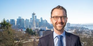 A white man in glasses and suit poses in from of the Seattle skyline