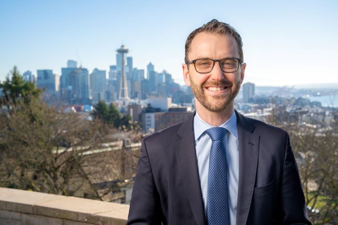 A white man in glasses and suit poses in from of the Seattle skyline