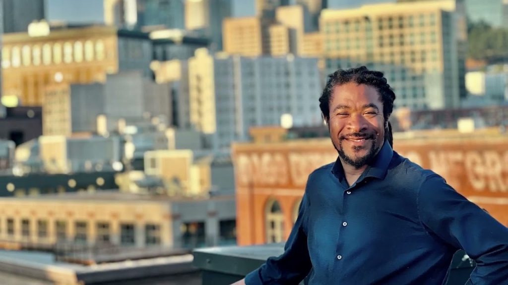 Rico is a Black man with braids and a button up shirt posing in front of the Seattle skyline