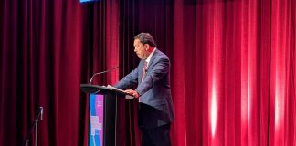 Harrell stands at a lectern and looks down as his notes. He wears a grey suit and stands in front of a red curtain.