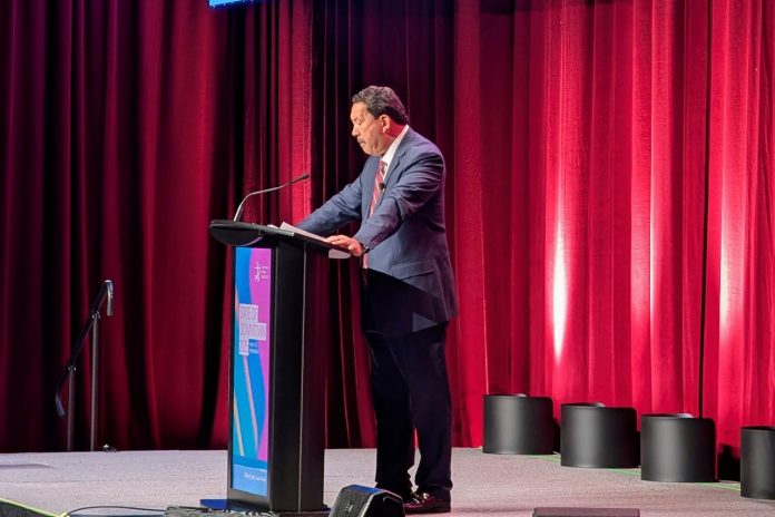 Harrell stands at a lectern and looks down as his notes. He wears a grey suit and stands in front of a red curtain.