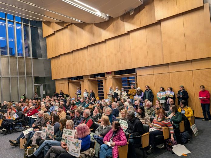 The Seattle council chambers are filled to the brim. People hold signs to promote their views.