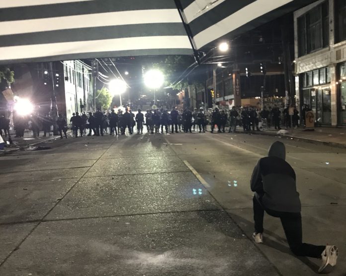 A protester stands just to the right of a gaggle of protesters. A line of police officers approach.