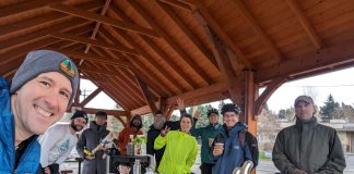 a group of 15 people under a picnic shelter around a picnic with coffee and pastries on it