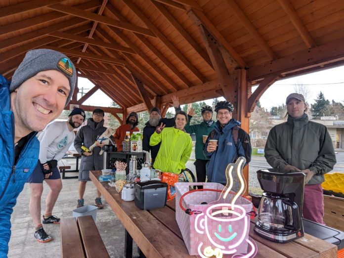 a group of 15 people under a picnic shelter around a picnic with coffee and pastries on it