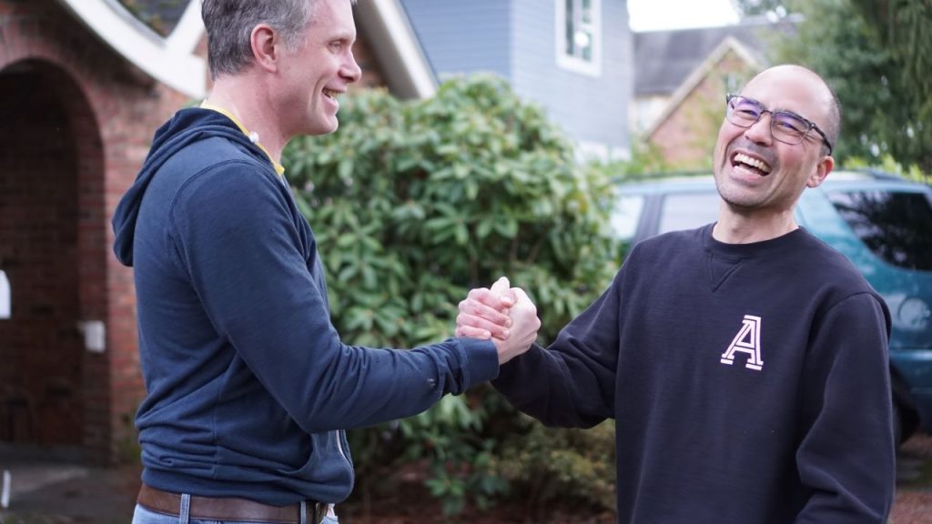 Eddie shakes hand with a man on the lawn in front of a brick house. He wears a big grin.