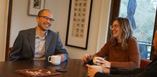 Lin is an Asian man with glasses and a suitcoat. He holds a coffee cup and sits next two people, one of them his wife.