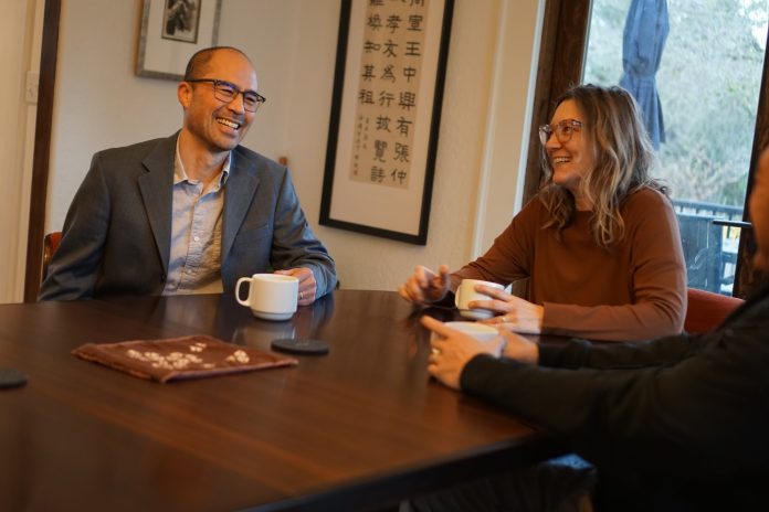 Lin is an Asian man with glasses and a suitcoat. He holds a coffee cup and sits next two people, one of them his wife.