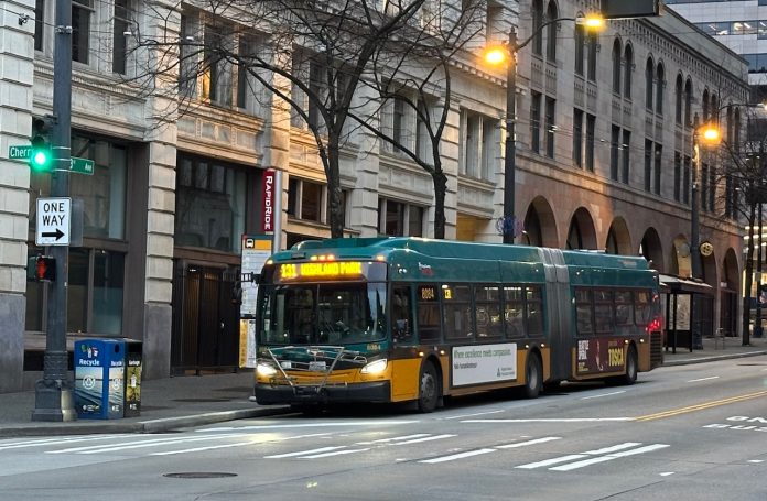 A bus sports King County Metro's green and gold livery with masonry buildings in the background.