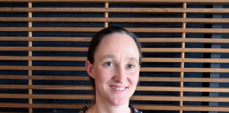 Katie wears a blue patterned blouse and stands in front of a wood slatted wall.