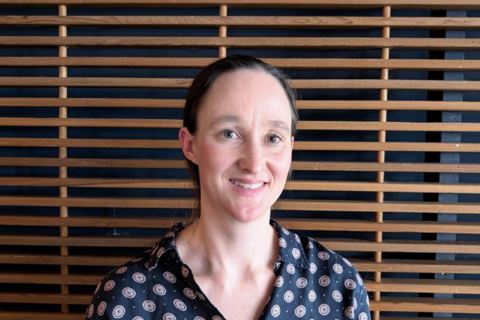 Katie wears a blue patterned blouse and stands in front of a wood slatted wall.