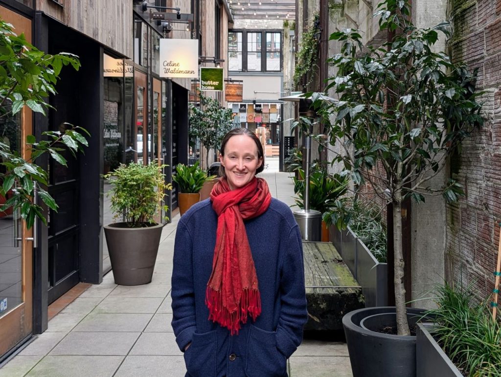 Wilson wear a red scarf and a dark blue coast and stand in the Chophouse Row alley which is lined by potted plants and restaurant storefronts.