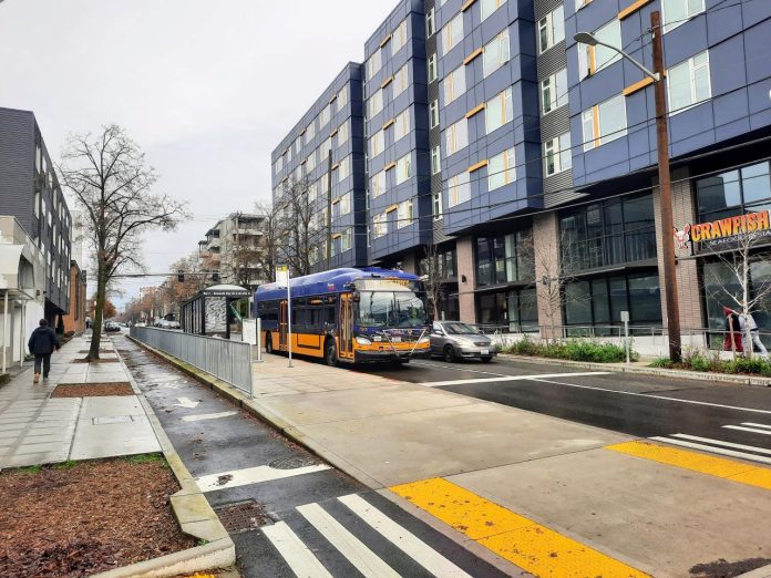 A travels down Roosevelt Way next to the protected bike lane. A large seven-story apartment building with a crawfish restaurant on the first floor is on the right.