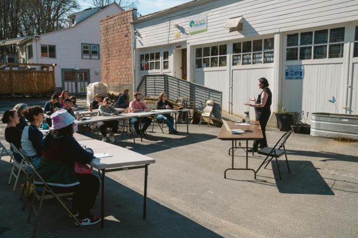 A woman leads a lass which is sitting around folding tables in the parking lot of the tool library building, and its rows of garage doors.