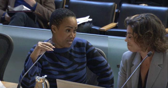 Washington is a black woman with short hair. She holds the microphone and looks to her left toward Natalie Walton-Anderson and the auditor as she delivers her rebuttal.